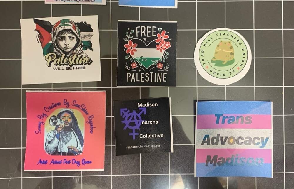 A cluster of Tessa's printed vinyl stickers on a cutting mat, including Free Palestine, Trans Advocacy Madison, a Madison Anarcha Collective mark, a Toad Hill Teachers Union badge, and an illustrated Sunny Ray Creations sticker.