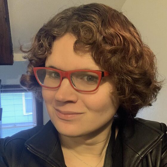 Tessa Price, a person with short curly auburn hair and red glasses, wearing a black leather jacket and smiling.